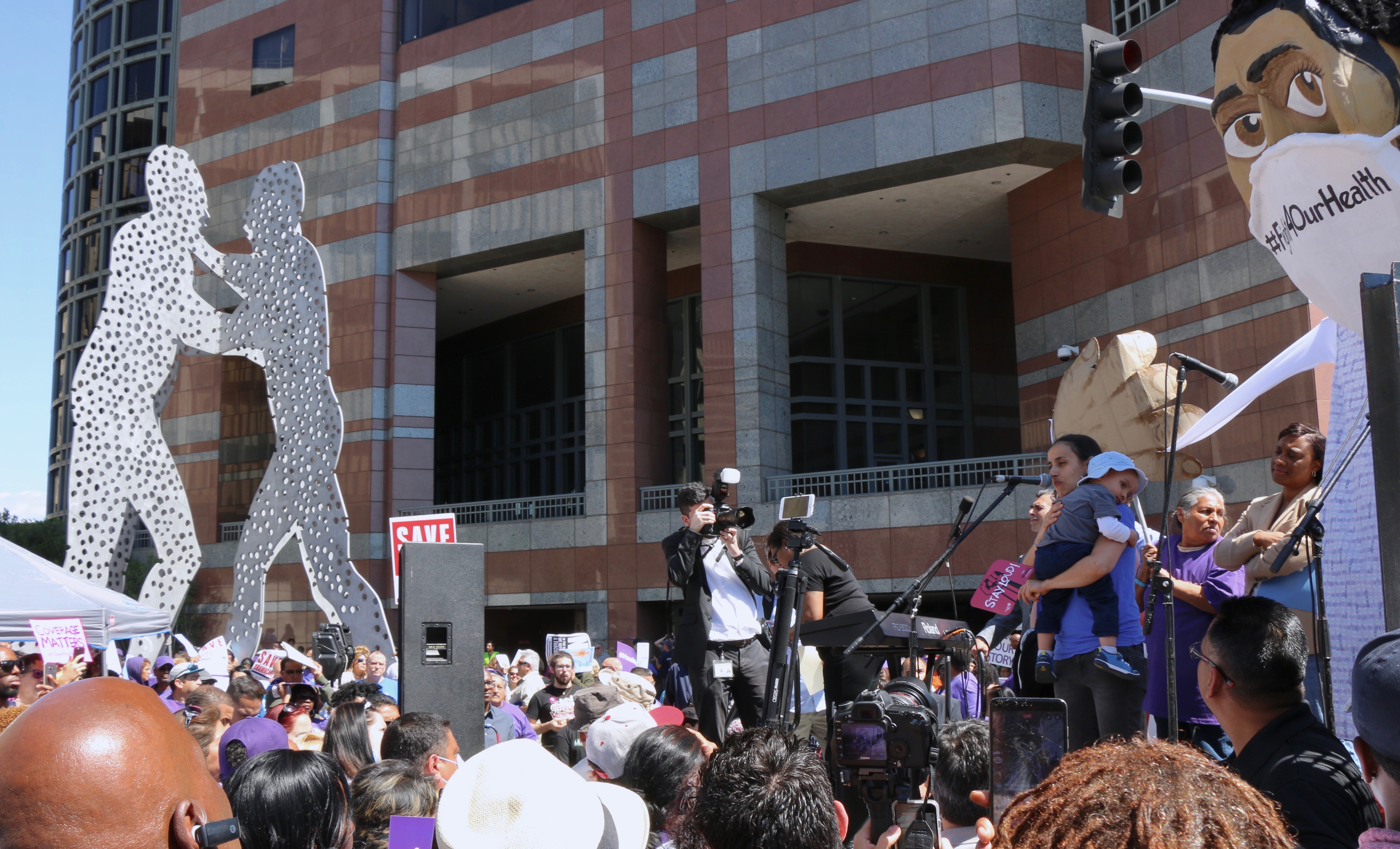 A mother helped by local Community Health Centers shares her story at the Save the Affordable Care Act march in Downtown Los Angeles, while Jonathan Borofsky's "Molecule Man" statue fights on.